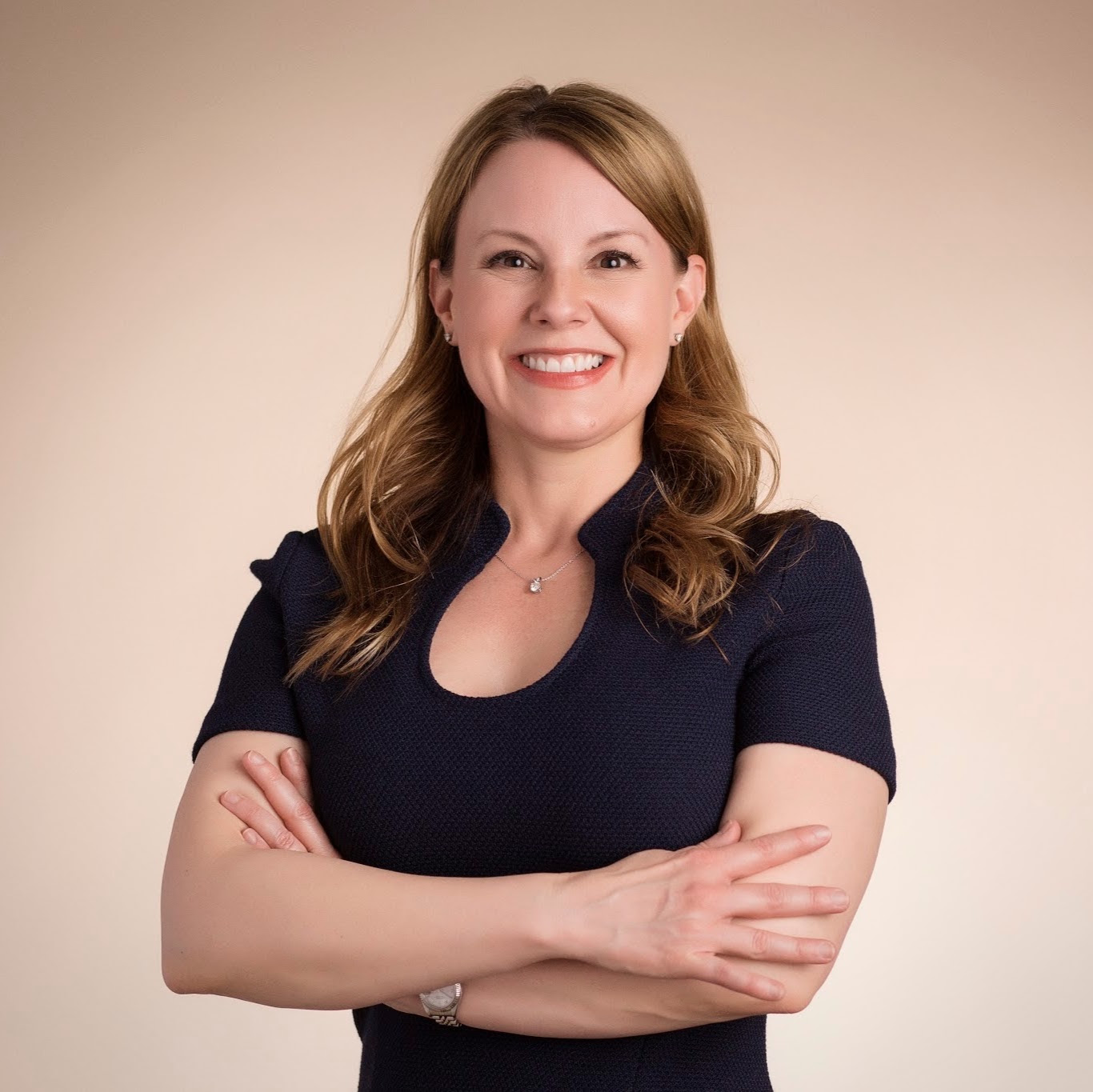 Woman with long brown hair smiling at camera, arms crossed, wearing dark blue top against neutral background